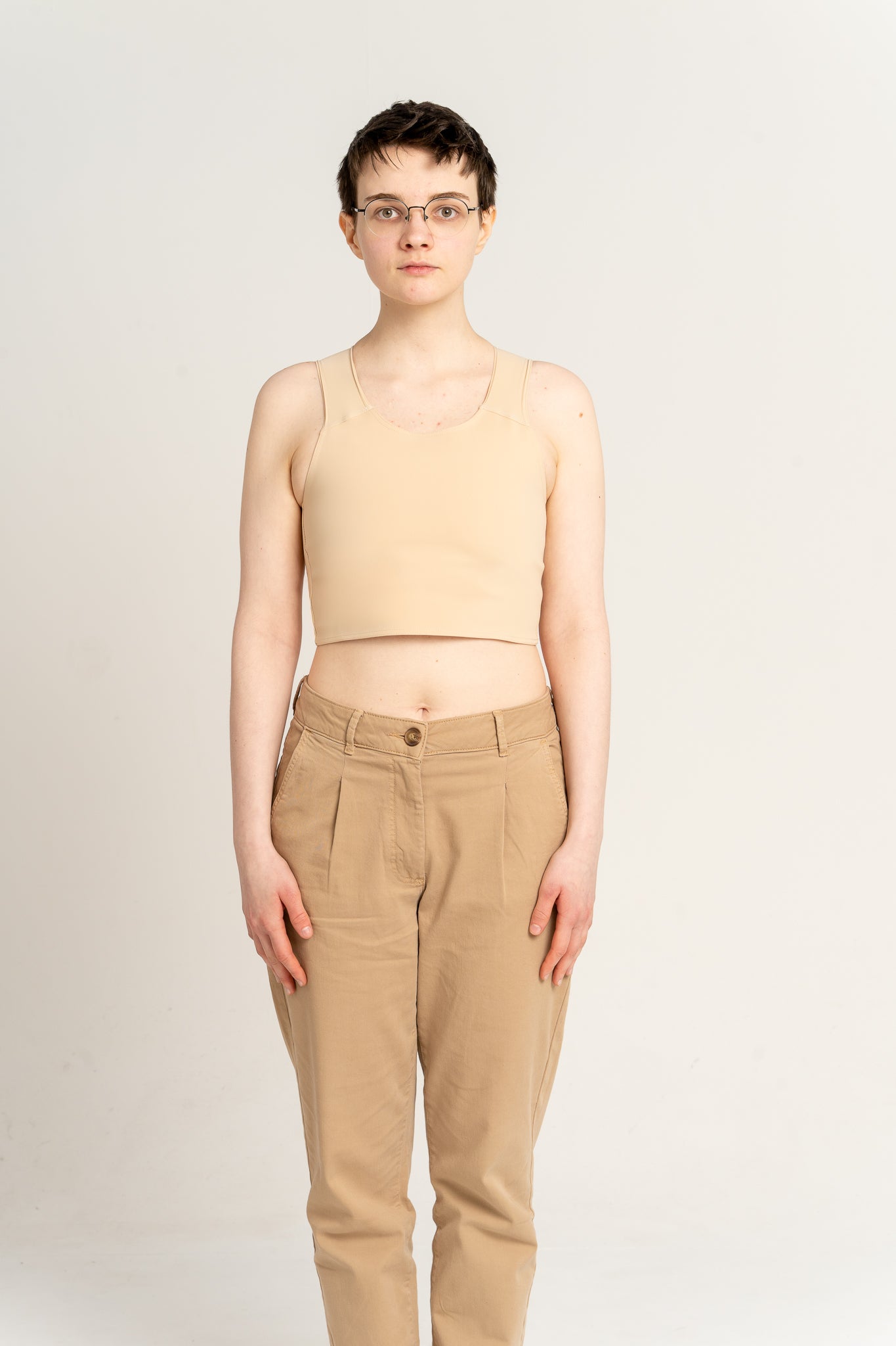 Person wearing a fair short chest binder and brown pants against a plain background