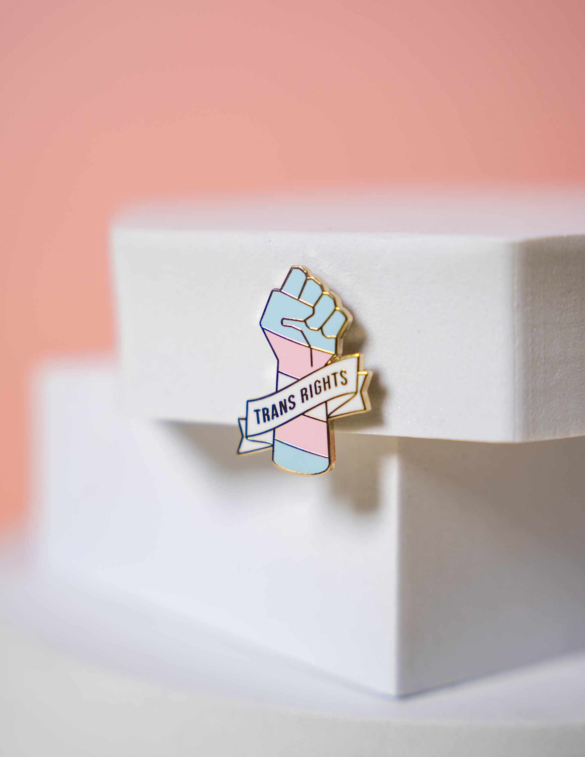 A trans flag enamel pin with a raised fist and a banner reading 'Trans Rights' on a white surface with a pink gradient background.