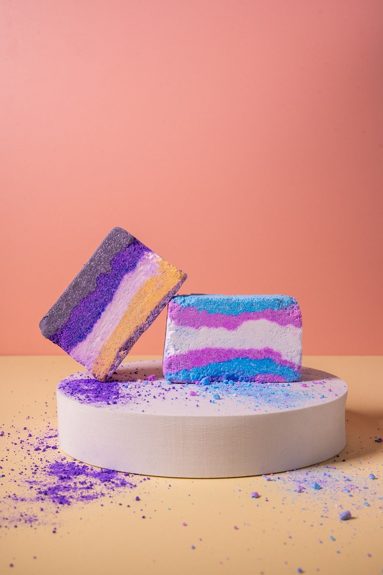 Colorful trans and non binary flag bath bombs on a white stand against a pink background