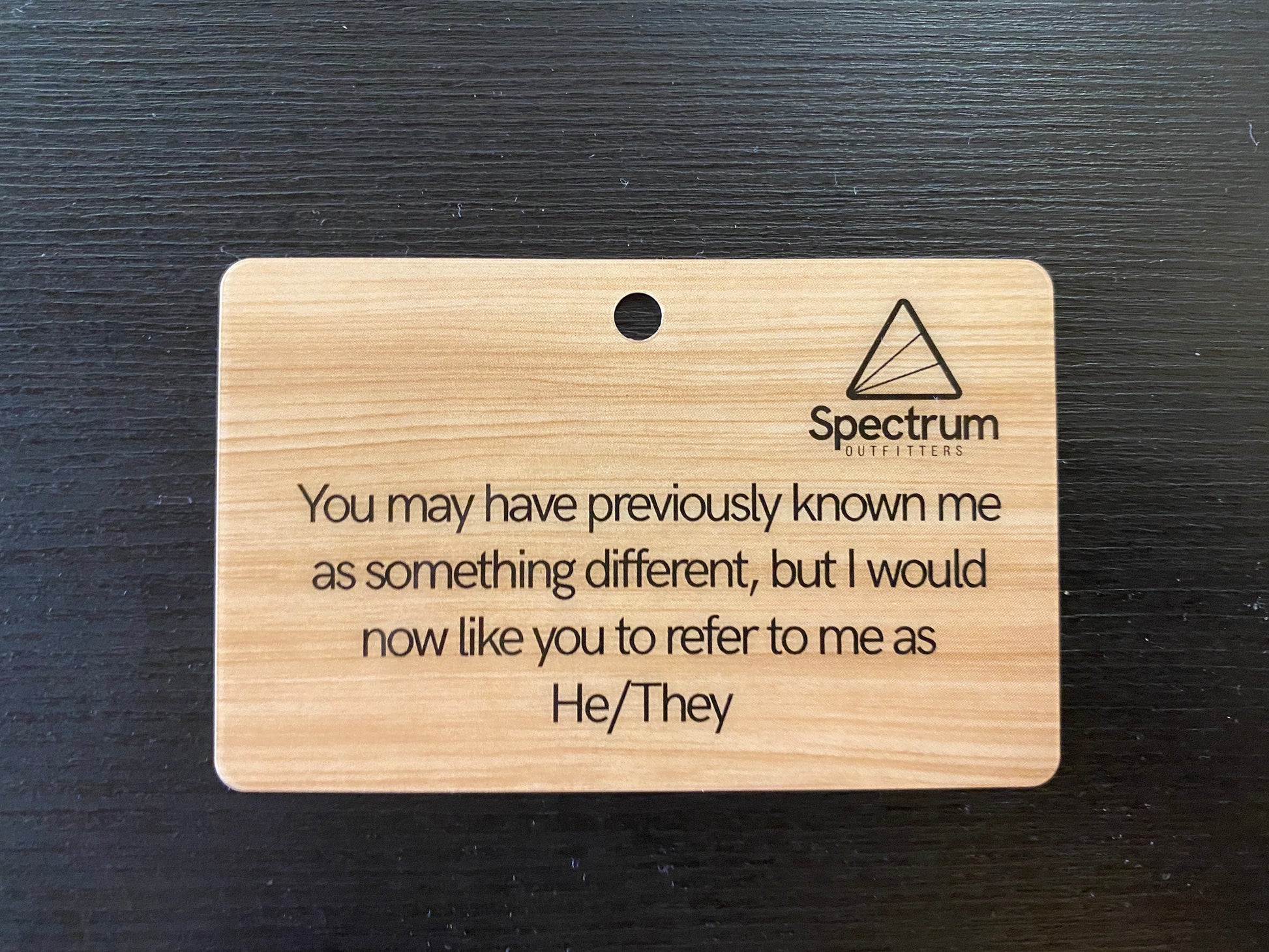 Wooden card with text reading 'You may have previously known me as something different, but i would now like you to refer to me as He/They' and a Spectrum Outfitters logo on a dark background