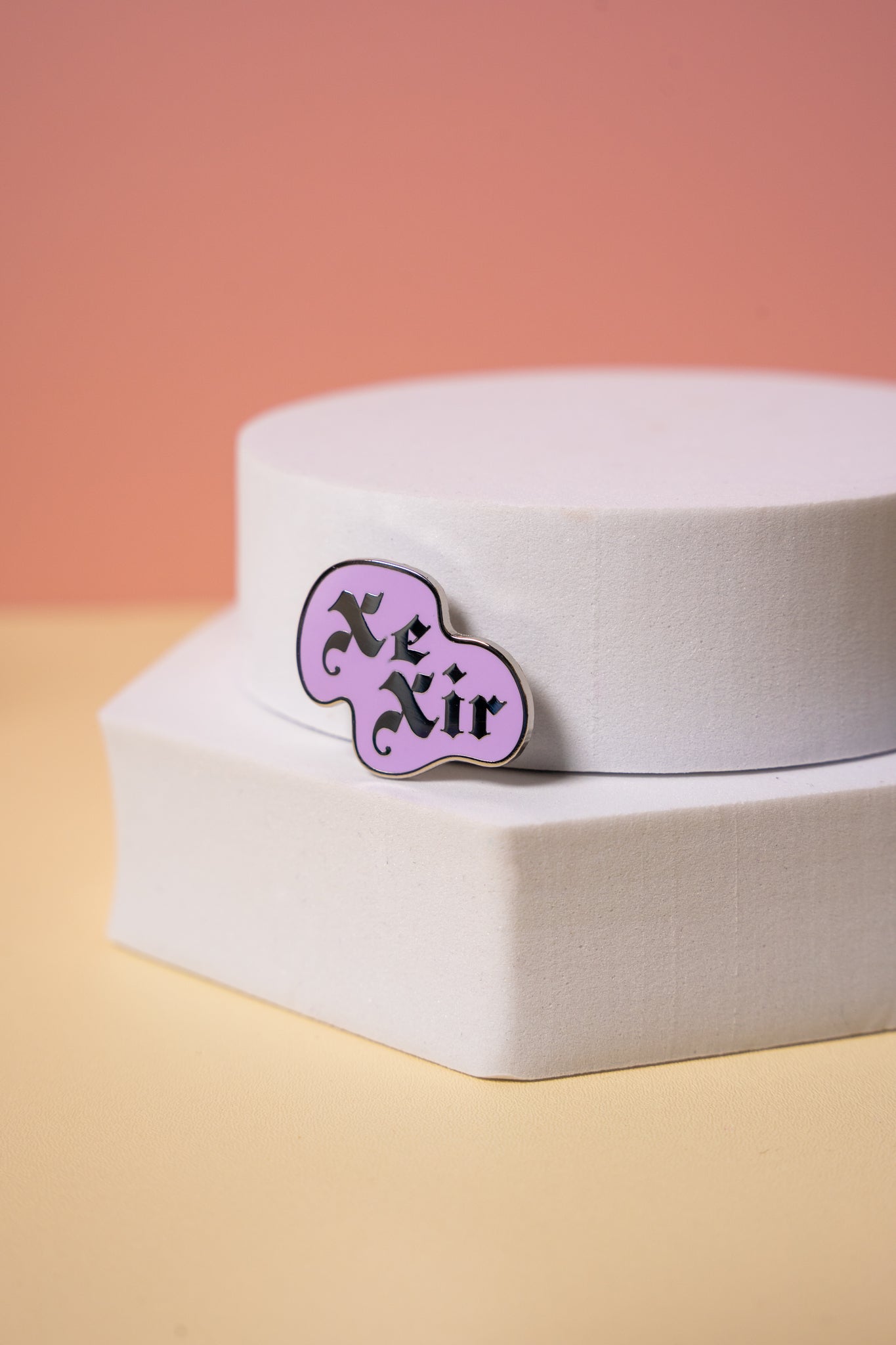 Purple 'Xe Xir' enamel pin on a white box with a pinkish-orange background