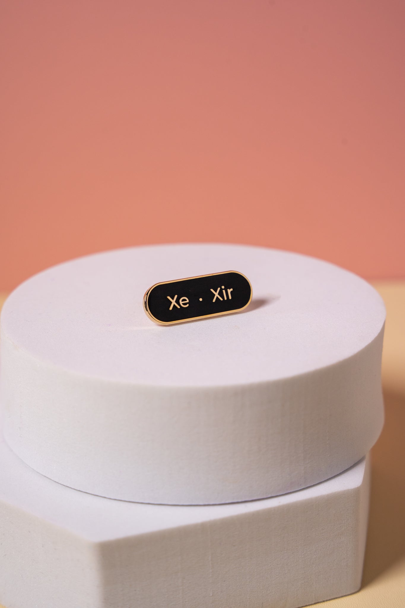 A gold and black enamel  'Xe Xir' pronoun pin on a peach background