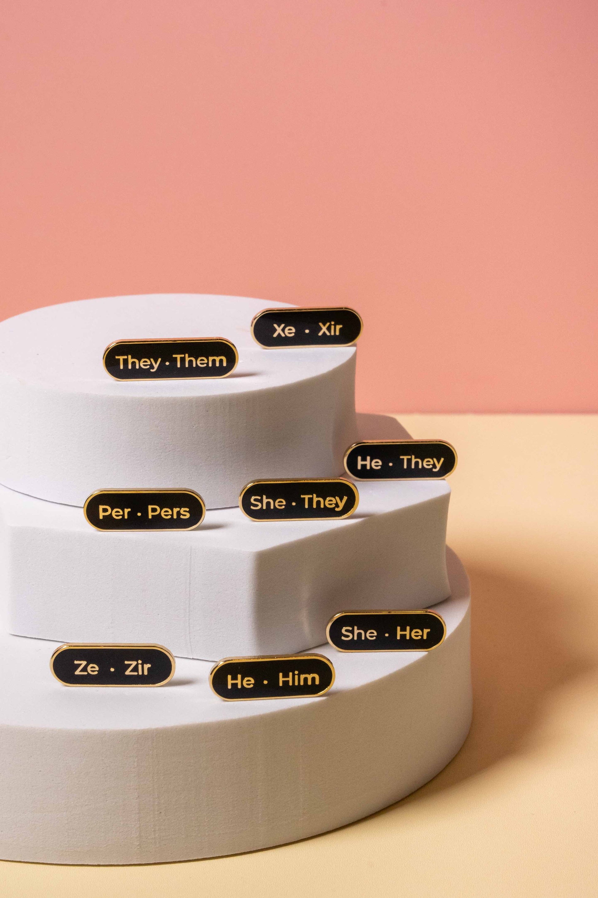 Assorted black and gold enamel pronoun pins displayed on a white stand.