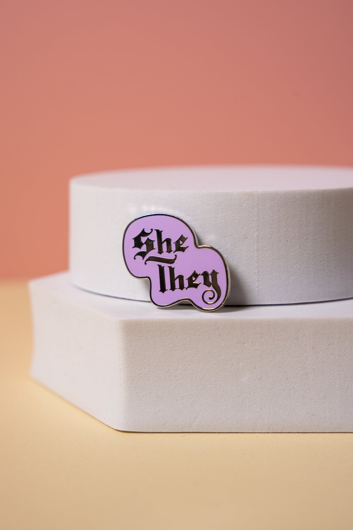 a purple enamel pin with text in gothic lettering showing the pronouns "she" and "they"
