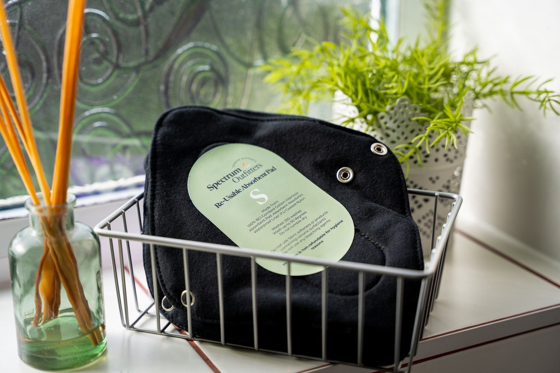Black reusable pads with a green label on a wire basket near a window with plants