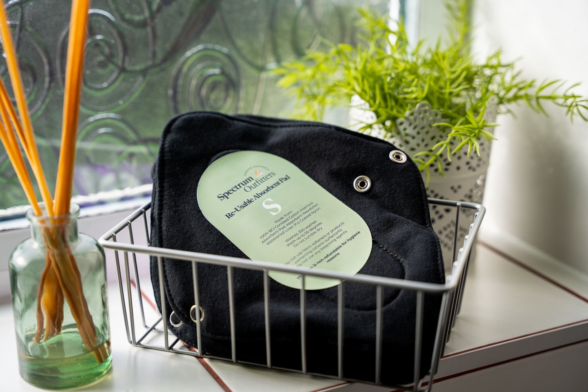 Black reusable pads with a green label on a wire basket near a window with plants